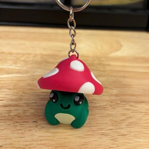 May include: A whimsical keychain featuring a green frog with a white belly and a red mushroom cap with white spots. The frog has a smiling face and large black eyes. The keychain is attached to a silver metal ring.