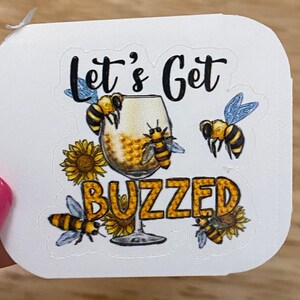 May include: White sticker with the text "Let's Get Buzzed" in black lettering. A glass of honey with bees and sunflowers are illustrated around the text.