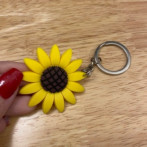 May include: A yellow sunflower keychain with a brown center. The sunflower is made of rubber and has a silver metal ring attached to it.