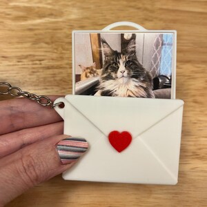 May include: A white keychain shaped like an envelope with a red heart. The envelope holds a small rectangular image of a cat. The keychain is attached to a silver chain.
