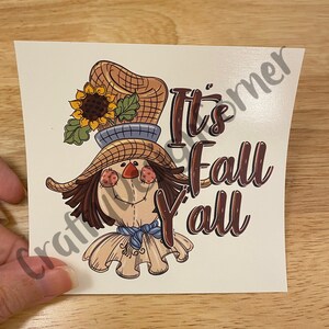 May include: A cute cartoon scarecrow with a sunflower in its hat and the text "It's Fall Y'all".