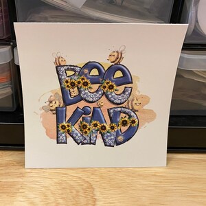 May include: A white card with a blue and yellow graphic that says "Bee Kind". The graphic features bees and flowers.