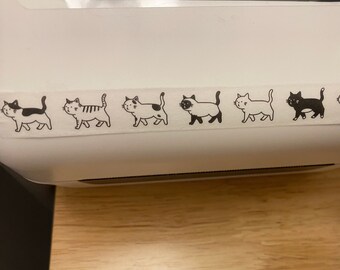 Puede incluir: Una impresora de etiquetas blanca con un rollo de cinta adhesiva de gatos negros y blancos. La cinta presenta un patrón repetitivo de gatos negros y blancos.