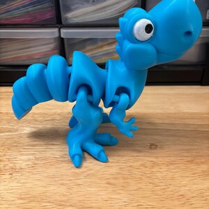 May include: A bright blue, articulated dinosaur toy. The toy has a segmented body, small arms, and large feet. The dinosaur's head features a large, white eye with a black pupil. The toy is made of plastic and sits on a wooden surface.
