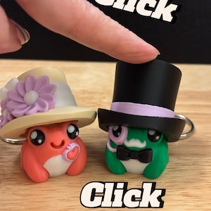 May include: Two whimsical frog keychains. One is red with a hat and flower, the other is green with a top hat and monocle. The word "Click" is written above and below the frogs.