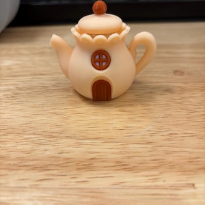Pode incluir: Uma chaleira caprichosa, cor de pêssego, com uma pequena tampa redonda encimada por uma esfera castanho-avermelhada. A chaleira apresenta uma porta castanha e uma janela redonda. A alça e o bico são curvos. A chaleira está sobre uma superfície de madeira.