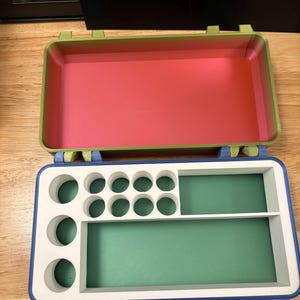 May include: A rectangular storage case with a hinged lid. The lid interior is a gradient of green to pink. The base features a blue outer frame with a white insert containing ten circular compartments and two rectangular storage sections.