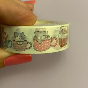 Pode incluir: Um rolo de fita washi com um padrão repetitivo de gatos em xícaras de chá. Os gatos estão em várias poses, incluindo dormir, sentar e olhar para cima. As xícaras de chá são decoradas com bolinhas e listras.