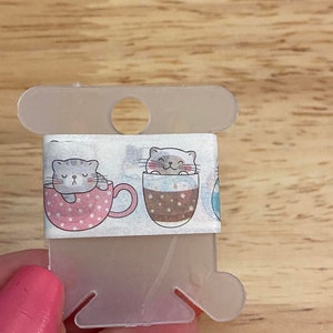 May include: Washi tape roll featuring a repeating pattern of cartoon cats in coffee cups. The cats are gray with closed eyes, and the cups are pink and brown with white polka dots. The tape is held on a clear plastic dispenser.