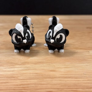 Può includere: Due piccole statuette di puzzola in bianco e nero con grandi occhi e una catena. Le puzzole hanno strisce bianche sulla schiena e dettagli bianchi sul viso e sui piedi. Sono su una superficie di legno.