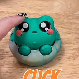 May include: A 3D printed green frog figurine with large black and white eyes and pink cheeks, seated inside a rounded, multi-colored base. The word CLICK is printed in orange letters at the bottom of the image.