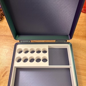 May include: An open, rectangular storage case with a colour-shifting iridescent exterior. The interior features a white insert with ten circular cutouts arranged in two rows, a rectangular compartment, and a larger open storage area at the bottom.