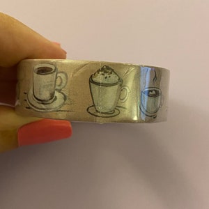 Puede incluir: Un rollo de cinta washi decorativa con un patrón repetido de tazas de café. Las tazas están ilustradas en un estilo vintage con colores marrón, blanco y negro.