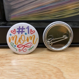May include: White button with a colorful design that says "#1 mom" with hearts and stars. The back of the button has a safety pin.