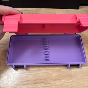 May include: A rectangular plastic storage case with a bright pink hinged lid and a purple base. The interior of the base features a row of five small, evenly spaced vertical dividers designed for holding slender items in place securely.