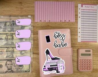 May include: A pink leather planner with a gold zipper and a clear plastic pocket on the front. The planner has a variety of stickers on it, including a black high heel shoe, a phone, and a coffee cup with the text "Boss Lady". The planner also has the text "Boss Babe" written on it in black cursive.