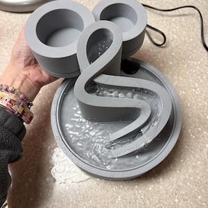 Puede incluir: Fuente de agua gris impresa en 3D con una base circular y un elemento de agua que fluye. La fuente tiene un diseño curvo único con agua cayendo en cascada hacia una cuenca. Dos recipientes circulares están unidos en la parte superior.