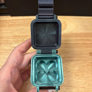 May include: A small, square, hinged container with a dark gray lid and a metallic teal base. The interior of the base features a four-leaf clover-shaped indentation with a central square detail. The lid has a textured, starburst-like pattern inside.