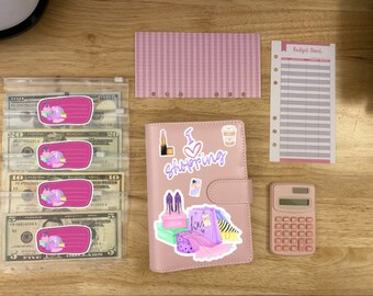 May include: A pink budget planner with stickers saying "I Shopping" and a pink calculator. There are also pink and white budget sheets, cash envelopes with money, and a pink striped divider.