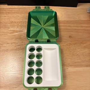 May include: A rectangular green plastic case with a hinged lid featuring a geometric star pattern on the interior. The base contains a white insert with ten circular holes arranged in two columns and a single rectangular mixing well for paint.