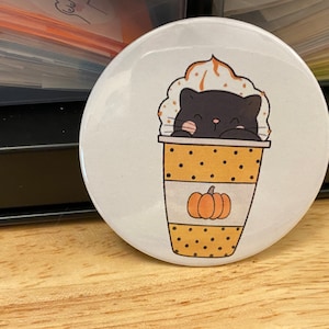 May include: A white button with a black cat peeking out of a yellow and white polka dot cup with whipped cream and a pumpkin on top.