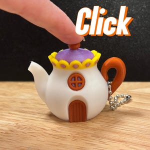 Pode incluir: Um chaveiro em forma de bule branco com tampa roxa, alça laranja e porta marrom. A tampa é adornada com detalhes amarelos e roxos. A palavra "Click" está escrita em letras laranja acima do bule.