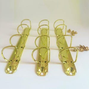 May include: Three gold-colored binder clips with three rings each, arranged diagonally. Small gold rivets are scattered nearby. The clips have a shiny, metallic finish, suitable for organizing documents.