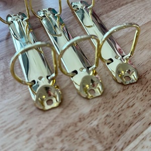 May include: Four gold binder clips with a D-ring design. The clips are arranged in a row on a wooden surface.