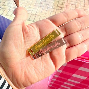 May include: Two rectangular metal tags with the word "Handmade" engraved on them. The top tag is gold and the bottom tag is rose gold.