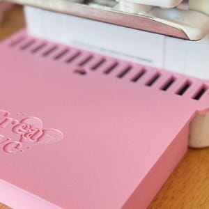 May include: A pink craft cutting mat with the embossed text "Dream Love" is positioned beneath a white die-cutting machine. The mat has a series of rectangular cutouts along the top edge. The items are on a wooden surface.