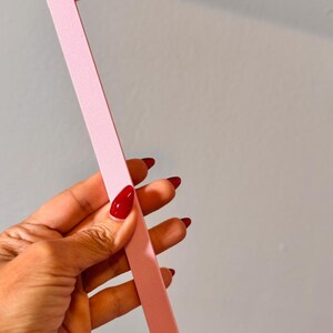 May include: A light pink L-shaped ruler or guide tool featuring a smooth matte finish. The tool has a small heart-shaped cutout at the end of each arm, providing a distinct visual detail on the otherwise straight, flat-edged structural design.