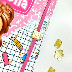 May include: Pink composition book with the word "Bella" on the cover. The book has a pink elastic band with a gold heart charm. There are three gold charms on a white surface: a flower charm with the words "I love Mom", a square charm with the words "I love Mama", and a square charm with a floral design.