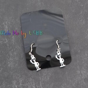 May include: A pair of silver dangle earrings with white alien figures. The earrings are displayed on a black background.