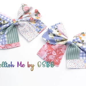 May include: Two handmade patchwork fabric hair bows in shades of blue, pink, and green with floral patterns. The bows are tied together with a white ribbon. "Embellish Me by OSBB" is written in rainbow colors below the bows.