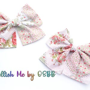 May include: Two large, white fabric bows with a patchwork design featuring pink and green floral patterns. The bows are tied with a knot in the center and have a slightly frayed edge. The text "Embellish Me by OSBB" is visible at the bottom of the image.