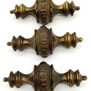 May include: Three antique-style brass drawer pulls arranged vertically. Each pull features a central bulbous section with embossed leaf-like detailing, flanked by tapered, multi-tiered ends that terminate in small, rounded points with a dark patina.