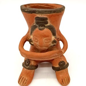 May include: A terracotta-colored clay vessel shaped like a seated figure with arms crossed over the chest. The figure features dark brown painted accents on the hair, eyes, limbs, and the rim of the wide, open-top container, showcasing a hand-molded form.