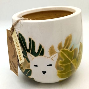 May include: A white ceramic planter featuring a relief design of a cat face surrounded by green and yellow tropical leaves. A brown paper tag attached with twine displays the text MADE BY HAND IN VIETNAM along with a map outline of the country.