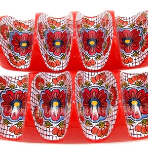 May include: Two red ceramic taco holders, each featuring four curved slots for holding tacos. The interior of each slot is decorated with a vibrant floral pattern in red, blue, and yellow, set against a white background with a fine grid design.