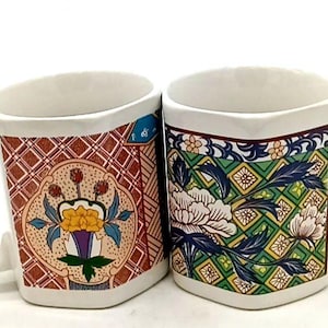 May include: Two white ceramic mugs with a hexagonal shape and handles. The left mug features a red grid pattern with a central floral motif. The right mug displays a green geometric background with large white flowers and blue leaves.