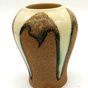 May include: A ceramic vase with a wide, rounded body and a narrow base. The surface features a textured, sandy brown finish contrasted by smooth, cream-colored glaze drips that cascade down from the rim, accented with dark green and brown edges.