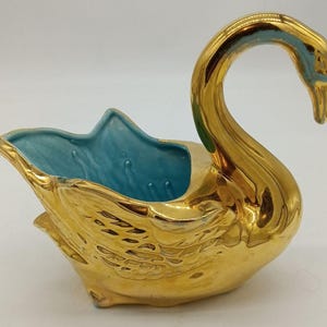 May include: A ceramic swan-shaped bowl featuring a reflective metallic gold exterior finish and a smooth, matte light blue interior. The swan is depicted with its neck curved downward, and the body displays molded feather details on the sides.