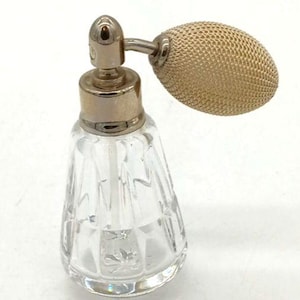 May include: A clear glass perfume bottle with a faceted geometric pattern on the body. The bottle features a metallic silver-toned collar and a pump mechanism attached to an oval-shaped, beige-colored fabric atomizer bulb with a fine mesh texture.