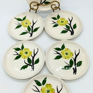 May include: A set of six off-white ceramic plates, each featuring a hand-painted design of a yellow flower with green leaves on a dark brown branch. One plate is displayed upright on a gold-toned metal stand, while five plates are arranged below.