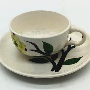 May include: A cream-colored ceramic teacup and saucer set featuring a hand-painted botanical design. The pattern displays a yellow flower with a red center, accompanied by dark green leaves and black branches extending across the cup and saucer.