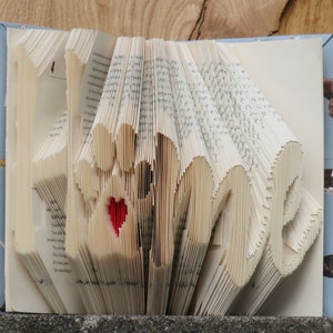 Peut inclure: Un livre dont les pages sont pliées en forme du mot "Home" avec un cœur rouge au centre. Le livre est relié en tissu bleu.