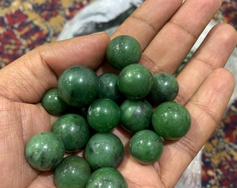 May include: A handful of polished, round, dark green gemstones. The stones are smooth and reflective, with variations in color and texture. The gemstones are held in an open hand, showcasing their natural beauty. The overall theme is gemstones.