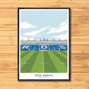 May include: A framed print of the RDS Arena, featuring a green field, blue seating, and a light blue sky with clouds. The stadium's name is displayed in white text at the bottom, with coordinates below.