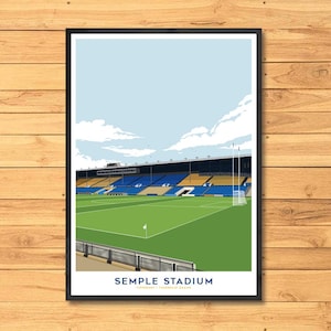 Op de afbeelding: Een ingelijste kunstafdruk van Semple Stadium, een sportlocatie, met een groen veld, blauwe en gele zitplaatsen en een lichtblauwe lucht met wolken. De naam van het stadion staat onderaan afgedrukt.