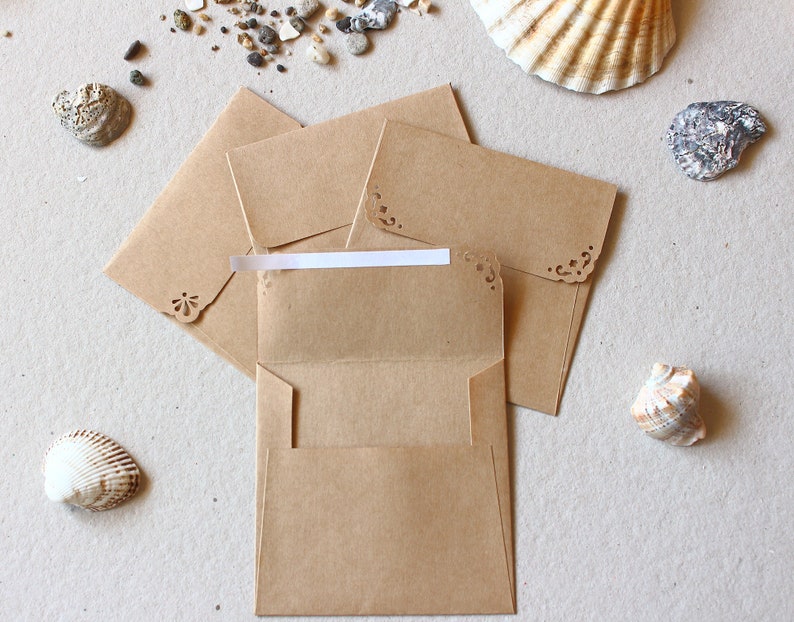 May include: Kraft paper envelopes with decorative corner cutouts, one open. The envelopes are arranged with seashells and pebbles on a neutral surface, creating a beach theme. Suitable for stationery or invitations.